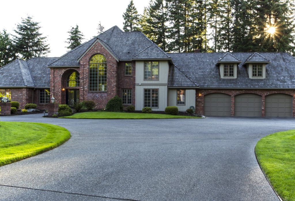 Driveway to large brick and cedar home with trees and sunlight in background